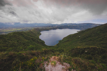 The image shows Cuicocha Lake, a stunning crater lake in Ecuador, nestled within a volcanic caldera. Deep blue waters reflect the sky, surrounded by steep, green hills and distant mountains under a cloudy skyの写真素材