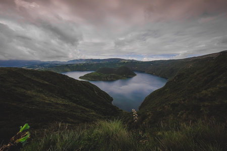 The image shows Cuicocha Lake, a stunning crater lake in Ecuador, nestled within a volcanic caldera. Deep blue waters reflect the sky, surrounded by steep, green hills and distant mountains under a cloudy skyの写真素材