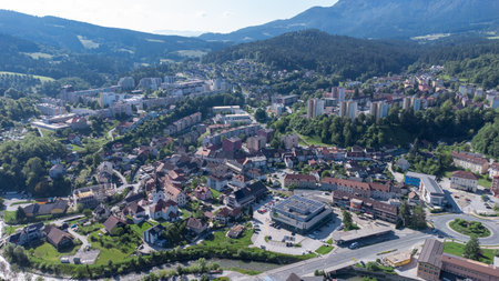 Aerial view of Ravne na Koro kem, a town nestled in a lush valley surrounded by rolling green hills and mountains under a partly cloudy sky. The town features a mix of residential areas, industrial buildings, and a prominent sports field with a running track. Green spaces and forests dominate the landscape, highlighting the areas natural beauty.の写真素材