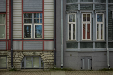 The image shows two adjacent buildings in Tallinn with distinct architectural styles. The left building features a red and gray facade with large windows and a stone base, while the right building has a gray exterior with ornate bay windows and a red interior glimpse, both accented by downspouts and a paved walkway.の写真素材