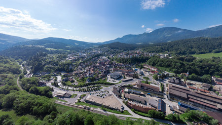 Aerial view of Ravne na Korokem, a town nestled in a lush valley surrounded by rolling green hills and mountains. The town features a mix of residential areas, industrial buildings, and a prominent sports field. Green spaces and forests dominate the landscape, highlighting the area's natural beauty.の写真素材