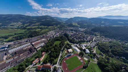 Aerial view of Ravne na Koro kem, a town nestled in a lush valley surrounded by rolling green hills and mountains under a partly cloudy sky. The town features a mix of residential areas, industrial buildings, and a prominent sports field with a running track. Green spaces and forests dominate the landscape, highlighting the areas natural beauty.の写真素材