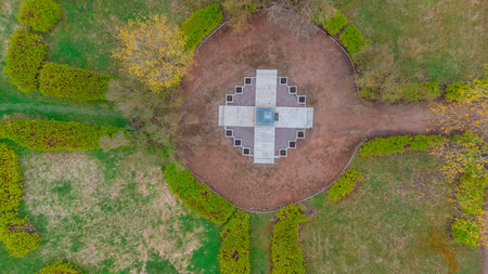 aerial view from Narva shows a circular garden with a cross-shaped monument at its center, and a curved road, Bare trees, capturing a serene autumn landscape., grassy fields, surrounded by neatly trimmed hedgesの写真素材
