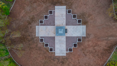 aerial view from Narva shows a circular garden with a cross-shaped monument at its center, and a curved road, Bare trees, capturing a serene autumn landscape., grassy fields, surrounded by neatly trimmed hedgesqの写真素材