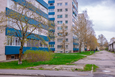 Narva residential area features multi-story brick buildings with blue accents, bare trees with early spring buds, and a grassy courtyard. A person walks a dog on a cracked road under a cloudy sky.の写真素材