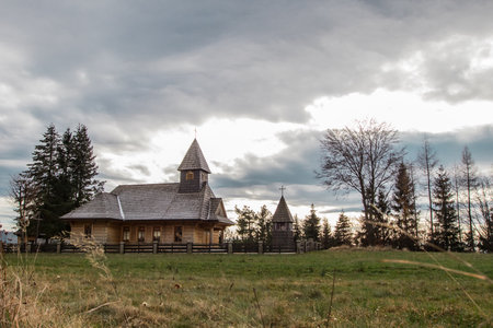A quaint wooden Church of Saint John the Baptist in Istebna, south of Wis a, nestled in the Beskidy mountains. Surrounded by lush grass, tall trees, and a cloudy sky, it features a shingled roof and modest tower, ideal for hikers exploring nearby trails.の写真素材