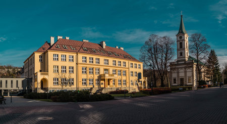 A vibrant yellow building with a red roof and a classic church with a green steeple in Wis as center, set against a clear blue sky. People walk on the cobblestone plaza, surrounded by trees, perfect for travel and urban imagery.の写真素材