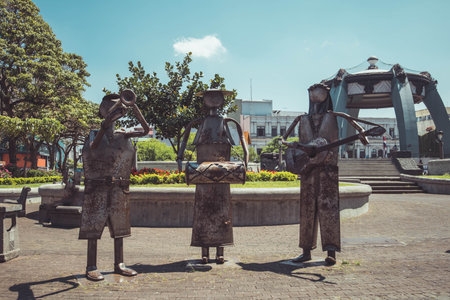 three metal sculptures of musicians in a park, playing a trumpet, drum, and guitar. They stand on a circular platform with greenery and flowers, under a clear blue sky. A pavilion and buildings are in the background, with a person sitting on a bench nearby.の写真素材