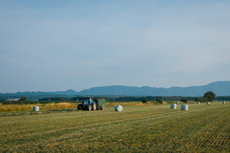 Vast field in Pragersko with a tractor baling hay into round bales under a clear blue sky. Rolling hills and distant mountains form a scenic backdrop, while the golden grass and green fields highlight a peaceful rural landscape.の写真素材