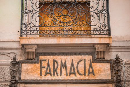 Vintage "Farmacia" sign in Udine, Italy, with ornate lettering on a marble plaque, flanked by wrought iron balcony and caduceus symbols, exuding historic charm.の写真素材