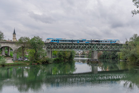 A modern blue passenger train crosses a steel railway bridge over the Krka River in Novo mesto, Slovenia. The church tower, lush trees, and river reflections create a peaceful scenic view.の写真素材