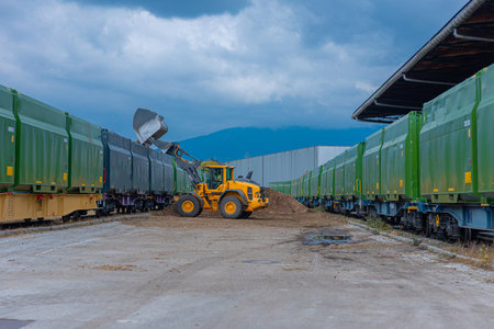A yellow loader unloads wood chips into green cargo train cars at a freight yard, with mountains and...