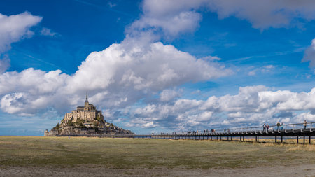 A long path leads across tidal fields to Mont Saint-Michel, the iconic island abbey in Normandy, France. The stone monastery rises dramatically under a wide, cloud-filled sky.の写真素材