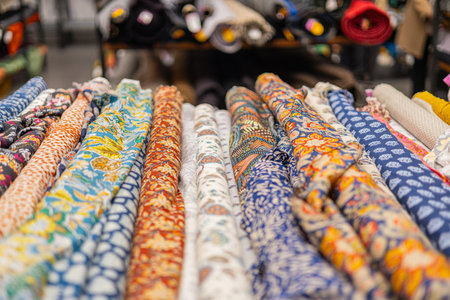 Close-up of neatly aligned rolled fabrics in a store, showcasing vibrant floral and abstract patterns in blues, reds, yellows, and whites. Blurred background hints at more textile rolls.の写真素材