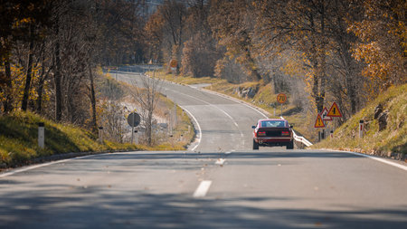 A classic red car drives down a winding, tree-lined road in autumn, with speed limit signs 50 and 70 and a sharp curve warning ahead, surrounded by golden foliage.の写真素材