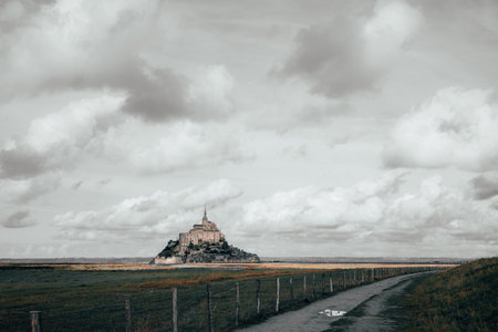 A long path leads across tidal fields to Mont Saint-Michel, the iconic island abbey in Normandy, France. The stone monastery rises dramatically under a wide, cloud-filled sky.の写真素材