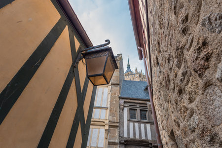 Narrow alley in Mont Saint-Michel, France. Half-timbered walls, vintage lantern, and stone facades frame a glimpse of the abbey s golden spire against a cloudy sky.の写真素材