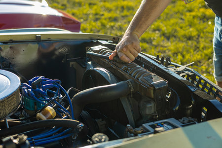 A close-up of a dirty hand working on a vintage car engine, gripping the radiator. Sunlight illuminates the blue wires, hoses, and mechanical parts in an outdoor grassy setting.の写真素材