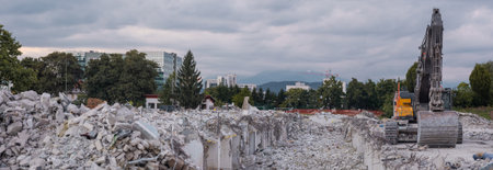 A demolition site filled with concrete rubble and twisted rebar. A large excavator stands ready amidst the debris, framed by urban office buildings and distant mountains under a cloudy sky in i ka.の写真素材