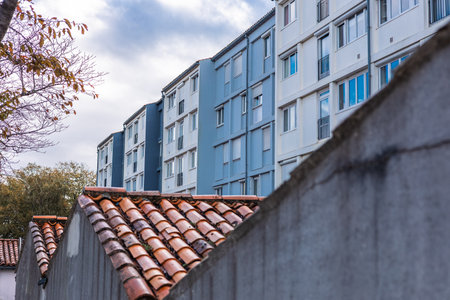 A visual contrast between modern residential blocks and traditional terracotta roof tiles framed by a concrete wall. The large pale blue and white buildings dominate the background under a cloudy sky.の写真素材