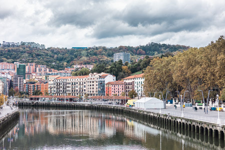 wide view of the Bilbao riverfront, with colorful buildings reflected in the calm water. The city rises up a wooded hillside under a dramatic, cloudy sky, showcasing urban and natural elements.の写真素材