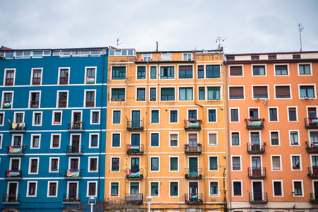 A symmetrical, full-frame view of three adjacent, brightly colored apartment buildings in Bilbao, featuring distinct blue, yellow-ochre, and orange facades with repetitive window and balcony patterns.の写真素材
