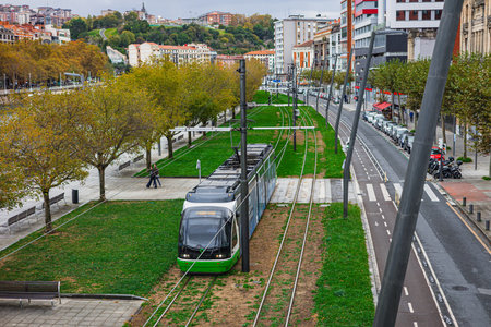An elevated view shows a green tram traveling along tracks embedded in a grassy median, flanked by a tree-lined promenade and a city street with parked cars and distant hillside buildings in Bilbao.の写真素材