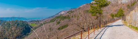 A panoramic view of a gravel cycle path along a rocky hillside with a rusty guardrail and a dark railway tunnel entrance among sparse, wooded trees.の写真素材
