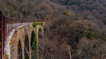 A panoramic view of a gravel cycle path along a rocky hillside with a rusty guardrail and a dark railway tunnel entrance among sparse, wooded trees.の写真素材