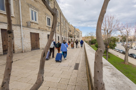 Tourists with luggage stroll along the historic Muraglia promenade in Bari Vecchia. Flanked by ancient stone facades and the Adriatic coastline, this elevated walkway offers scenic views of the sea.の写真素材