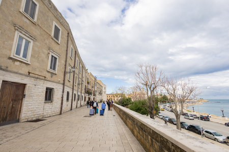 Tourists with luggage stroll along the historic Muraglia promenade in Bari Vecchia. Flanked by ancient stone facades and the Adriatic coastline, this elevated walkway offers scenic views of the sea.の写真素材
