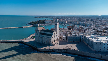 A panoramic coastal view of Trani shows the famous seaside cathedral overlooking turquoise waters, a long breakwater with lighthouses, and the historic old town bathed in bright Mediterranean sunlight.の写真素材