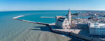 A panoramic coastal view of Trani shows the famous seaside cathedral overlooking turquoise waters, a long breakwater with lighthouses, and the historic old town bathed in bright Mediterranean sunlight.の写真素材