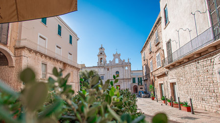A charming stone street in Trani leads to an ornate historic church, framed by old buildings, bright shutters, and sunlit Mediterranean colors, creating a warm and inviting southern Italian atmosphere.の写真素材