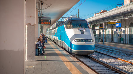 A sleek blue Intercity train stands at the platform in Trani Centrale station, Italy. The sunny railway scene features overhead lines, metal station canopies, and carriages waiting for passengers.の写真素材