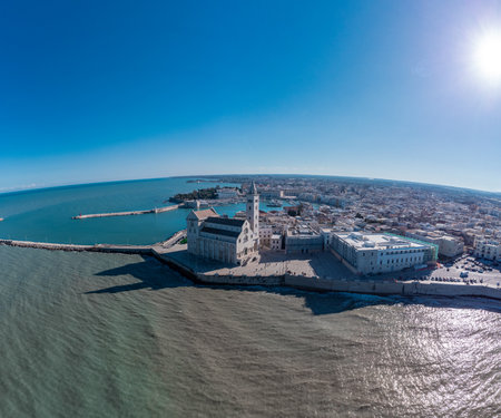 A panoramic coastal view of Trani shows the famous seaside cathedral overlooking turquoise waters, a long breakwater with lighthouses, and the historic old town bathed in bright Mediterranean sunlight.の写真素材