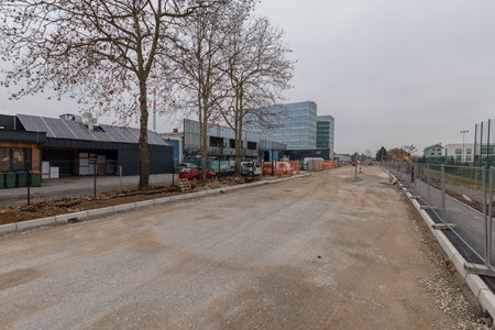 Active construction site on Magistrova street, Ljubljana, featuring an orange excavator, deep trench with concrete walls, workers laying utility pipes, and residential buildings under a cloudy sky.の写真素材