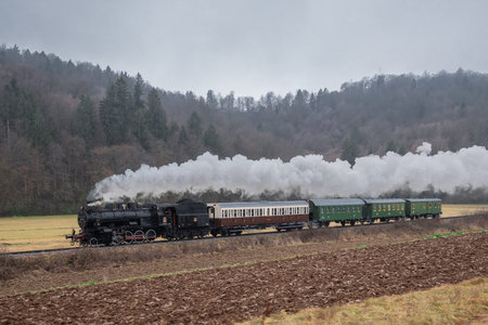 A vintage steam locomotive pulls classic passenger carriages through a quiet rural valley, releasing...
