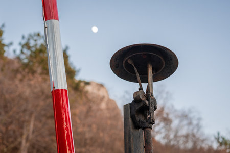 Bell detail on traditional red-and-white railway crossing barrier stands raised beside a quiet rural road. Weathered metal mechanics and chains contrast with bare autumn trees and earthy hills in the background.の写真素材