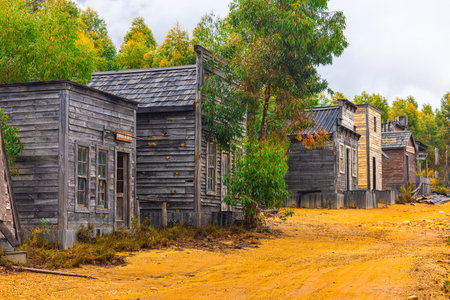 A row of weathered wooden buildings styled like an old Western frontier town, with signs for "Jail" and "Laundry," on a dusty dirt road in Somerset West. Trees partially obscure the structures, cyclists pass by, under a cloudy sky with autumnal foliage.の写真素材