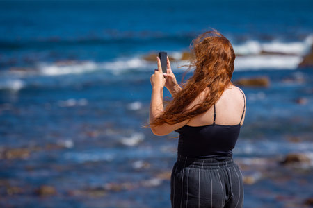 A woman stands by the rocky shoreline at the Cape of Good Hope, photographing rolling Atlantic waves...