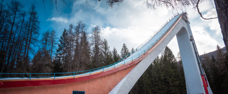 Ski jump ramp rising above alpine forest in Cortina d Ampezzo, Italy, with dramatic concrete structure, blue sky, winter light, and towering pines creating a striking mountain sports scene.の写真素材