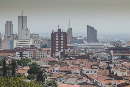 A panoramic view of Cali, Colombia, contrasting the red-tiled roofs of the historic district with the towering skyscrapers of the modern city center under a hazy, overcast sky.の写真素材