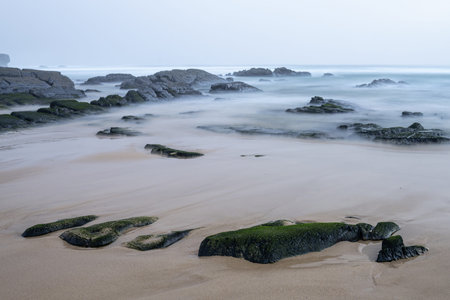 A tranquil scene at Amoreira Beach in Portugal, featuring mist-covered rocks and soft waves gently...