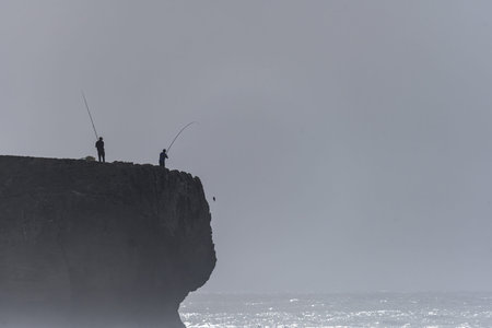 Silhouetted fishermen on the edge of the cliffs at Amoreira Beach, Portugal, casting their lines into the ocean below. A misty, atmospheric scene with a rugged coastline.の写真素材