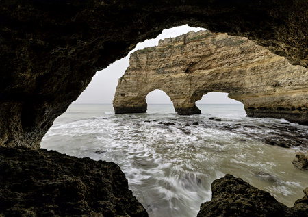A rocky outcrop at Marinha Beach, Portugal, silhouetted against the soft light of a cloudy sky. Waves gently lap at the shore, creating a serene coastal scene.の写真素材