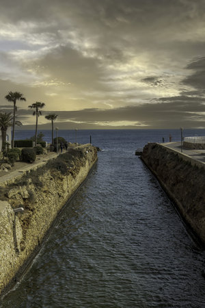 Scenic coastal walkway at Cala en Bosch, Menorca, flanked by palm trees and white buildings. A canal leads to the open sea under a partly cloudy sky.の写真素材