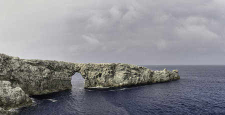 Stunning view of Pont de Gil in Menorca, a natural rock arch extending into the deep blue sea under an overcast sky. A magnificent coastal formation.の写真素材