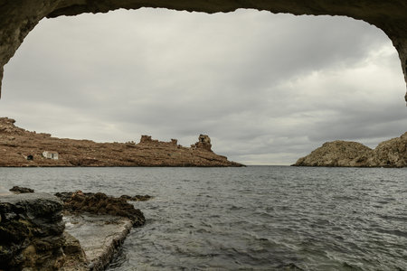 Dramatic view from inside a coastal cave at Cala Morell, Menorca, showing rugged rocky formations and crashing waves under a cloudy sky.の写真素材