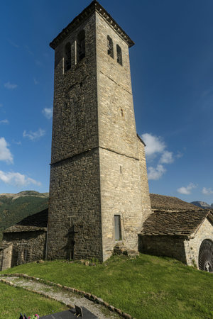 A historic stone church with a bell tower, surrounded by greenery and set against a bright blue sky and mountainous backdrop in Fanlo.の写真素材